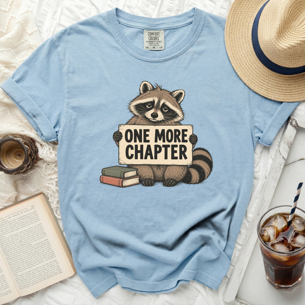 Light blue t-shirt with raccoon holding "ONE MORE CHAPTER" sign and books.