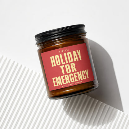 Amber candle in glass jar with red label reading "Status: Holiday TBR Emergency"