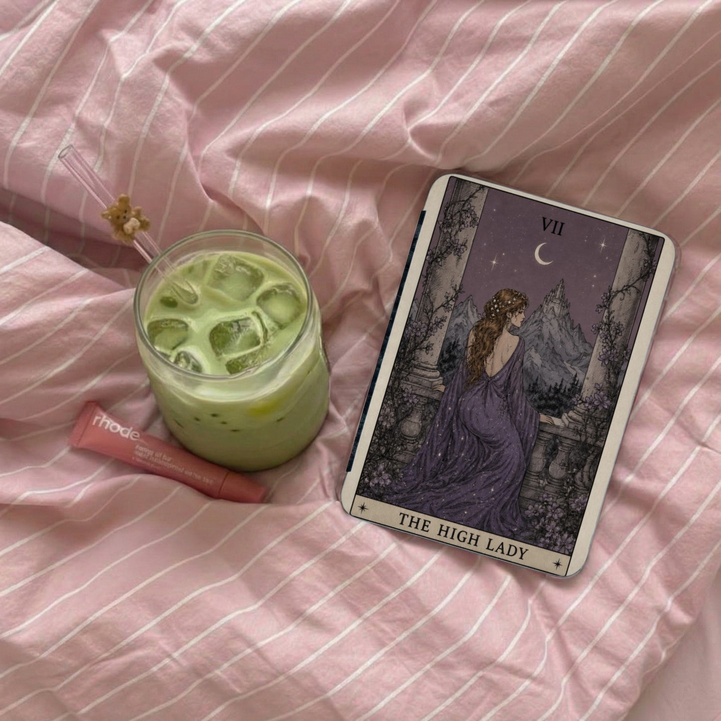 Tarot card "The High Lady" with a green iced drink and lip balm on pink striped sheets.