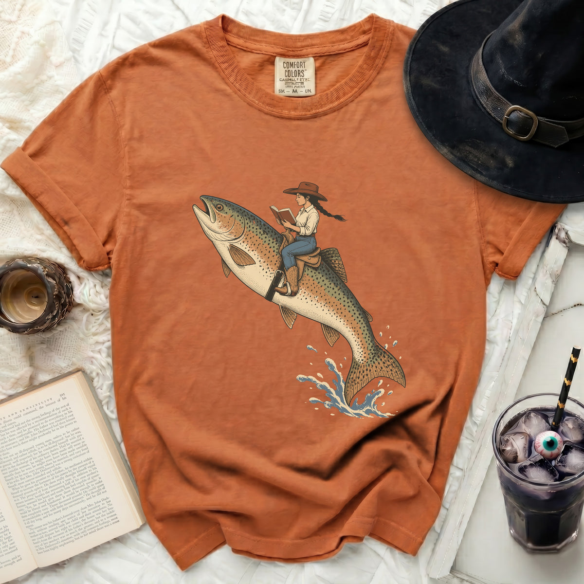 Orange t-shirt with illustration of a woman riding a trout and reading a book.