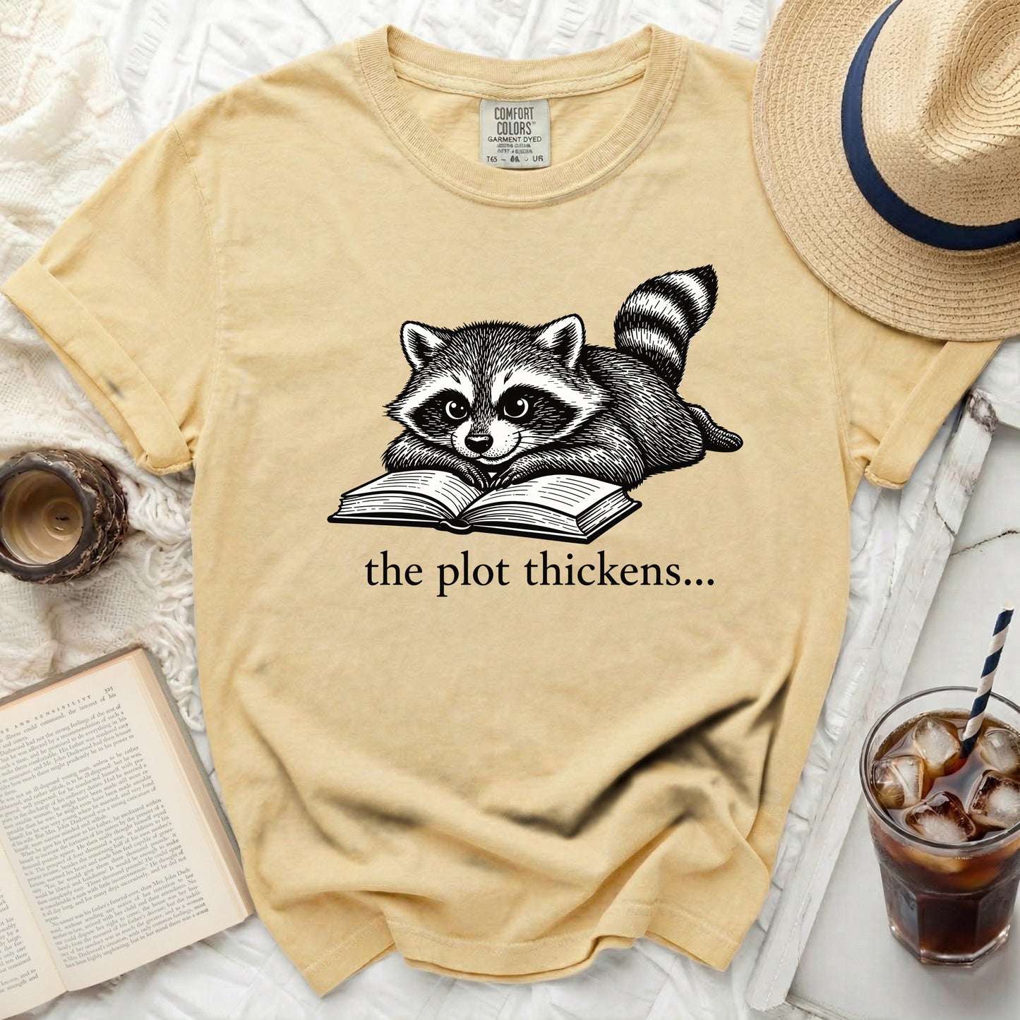 Yellow t-shirt with a drawing of a raccoon reading a book, text reads "the plot thickens...".