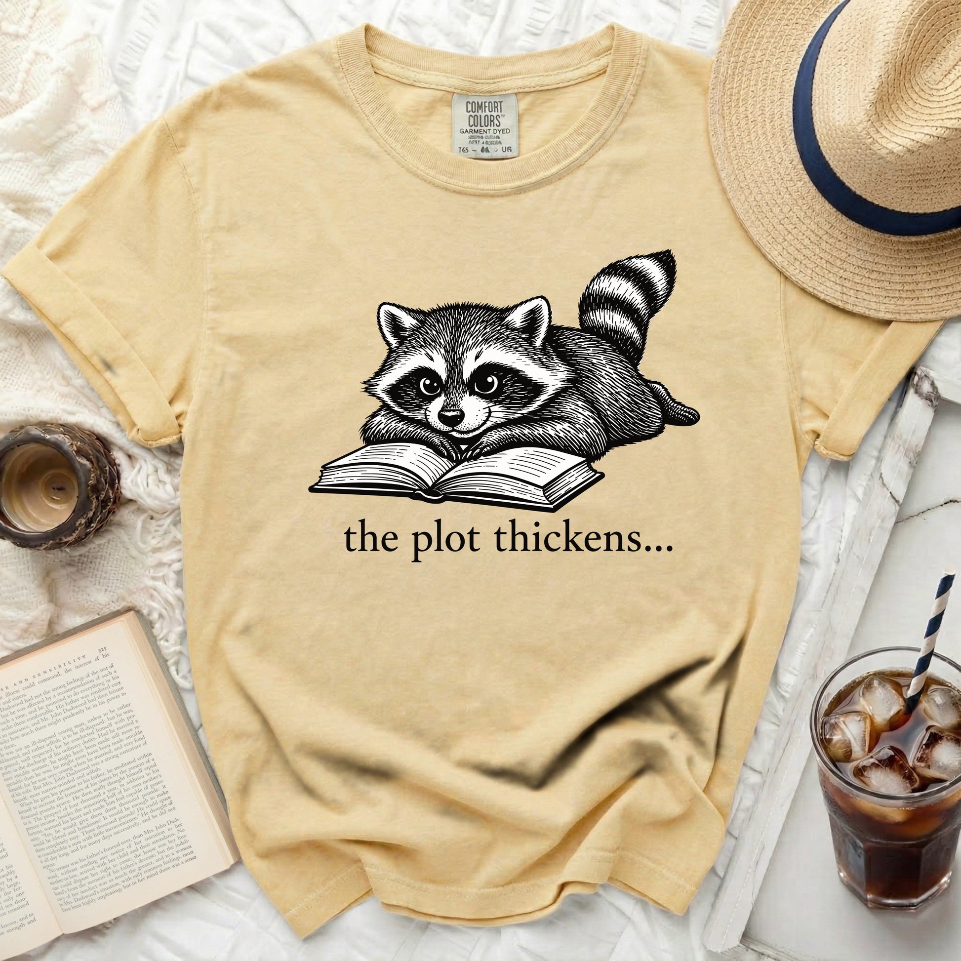 Yellow t-shirt with a drawing of a raccoon reading a book, text reads "the plot thickens...".