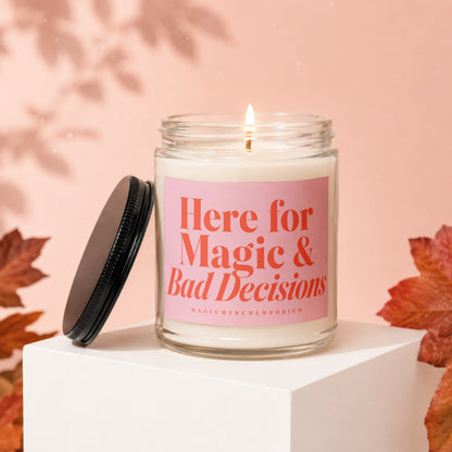 Glass candle with lit wick and pink label reading "Here for Magic & Bad Decisions".