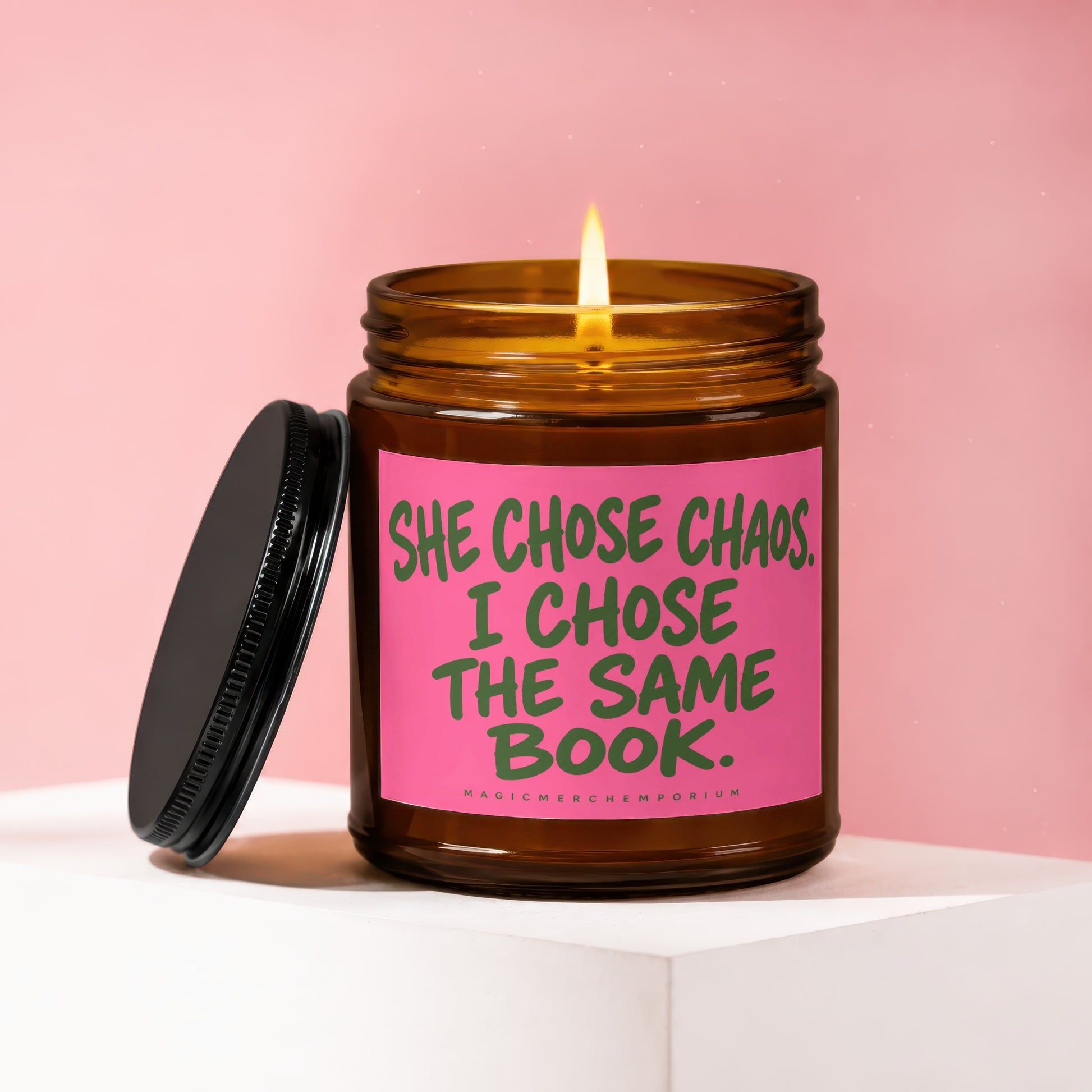 Amber jar candle with lit flame and black lid. Pink label reads "SHE CHOSE CHAOS. I CHOSE THE SAME BOOK."