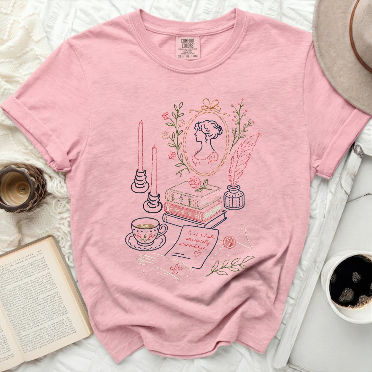 Pink t-shirt with embroidered literary scene including portrait, books, tea cup, quill, and text.