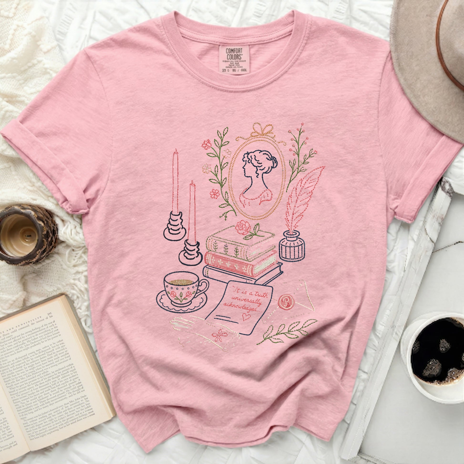Pink t-shirt with embroidered literary scene including portrait, books, tea cup, quill, and text.