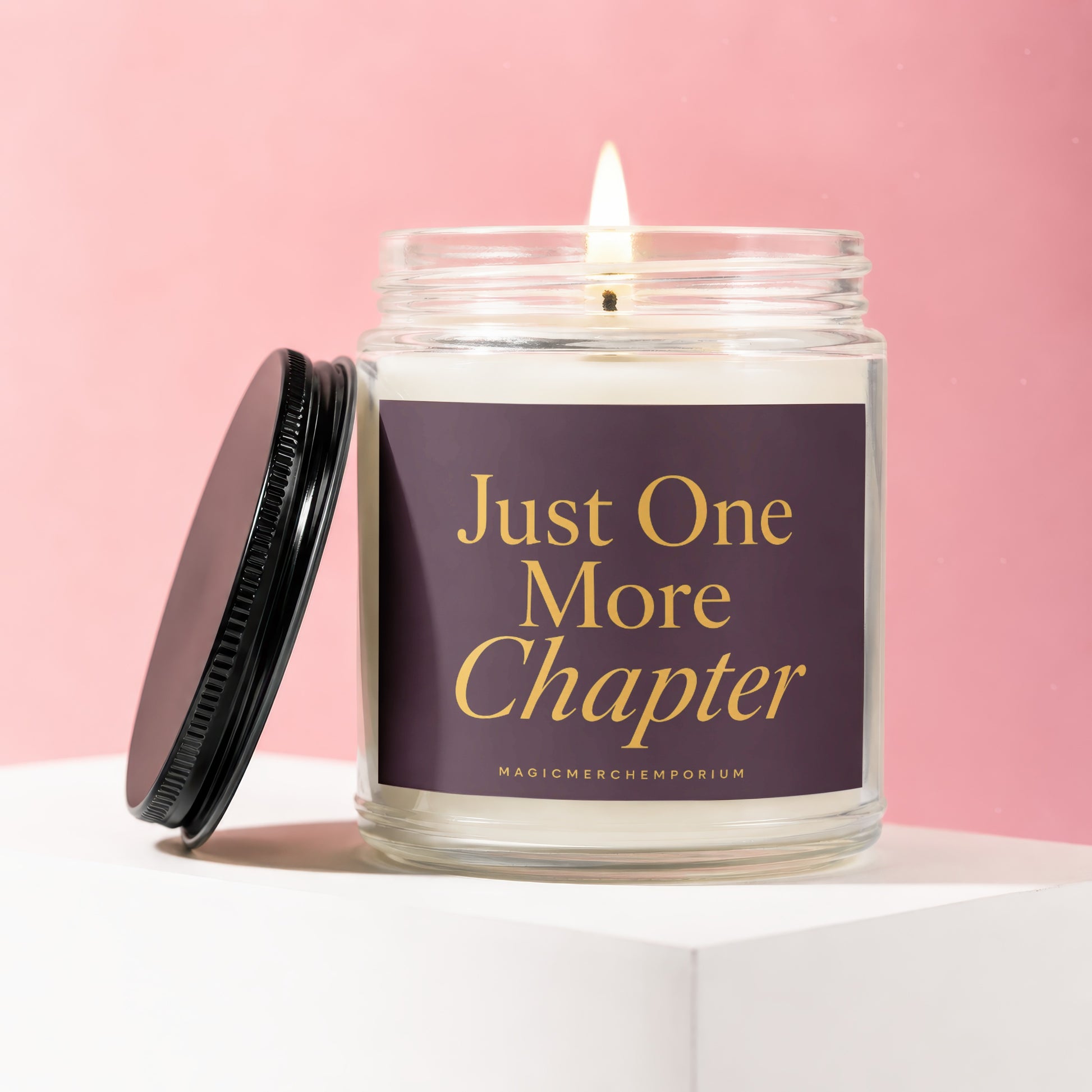Glass candle with lit wick, black lid nearby, reads "Just One More Chapter".