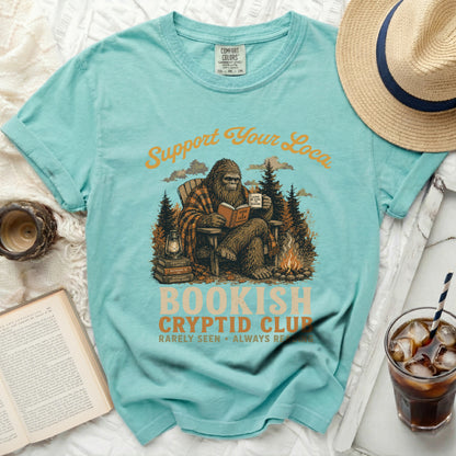 Bigfoot reading a book and mug with "Support Your Local Bookish Cryptid Club" text.