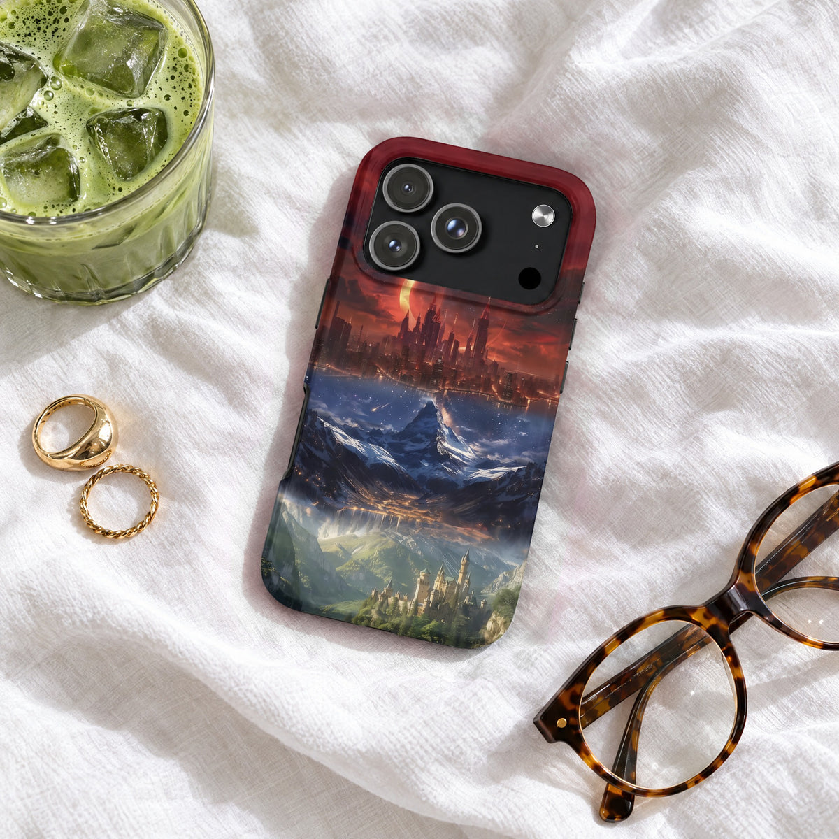 A smartphone case featuring a fantasy landscape design lies on a textured white sheet beside glasses, rings, and iced green tea.
