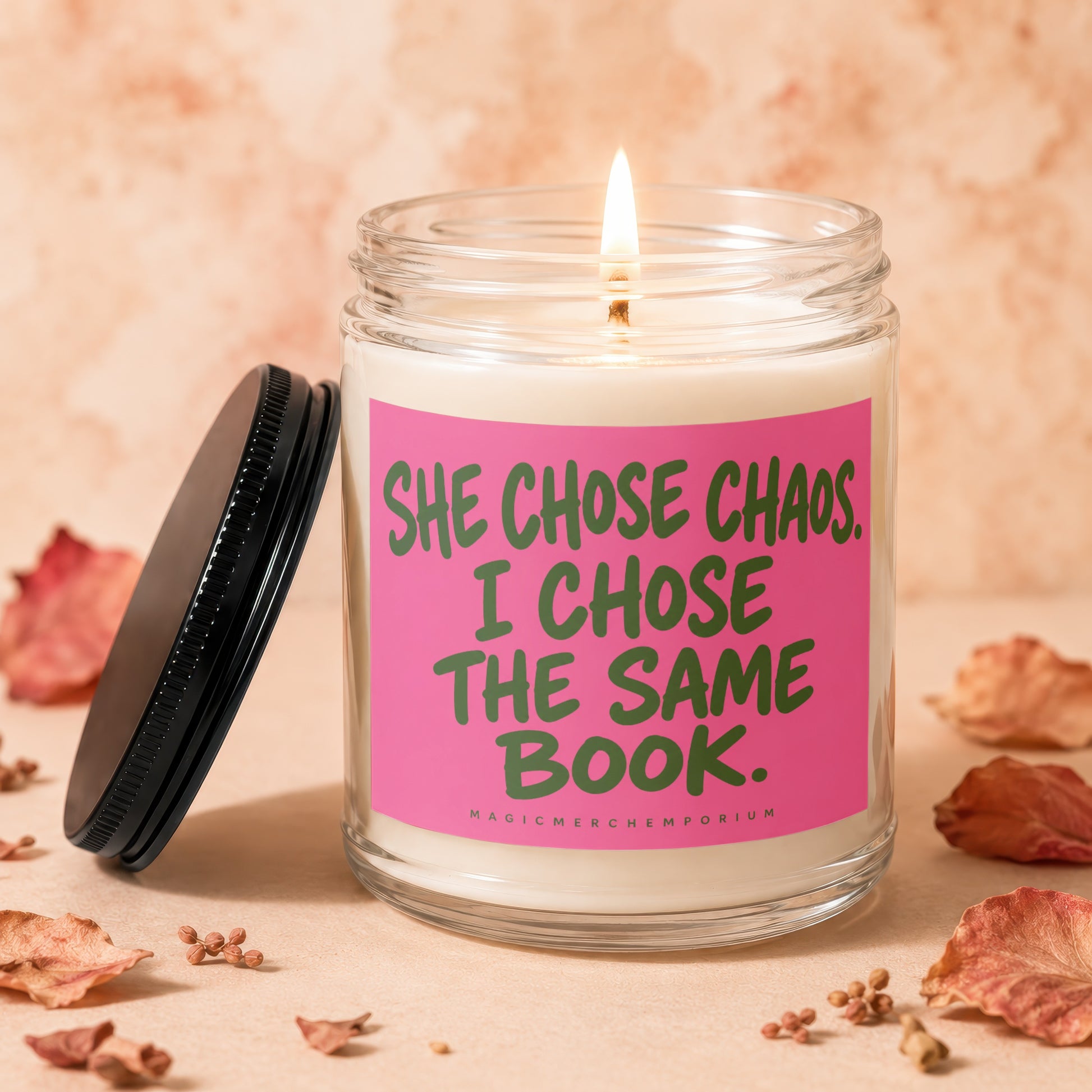 Lit glass candle with pink label reading "She chose chaos, I chose the same book."