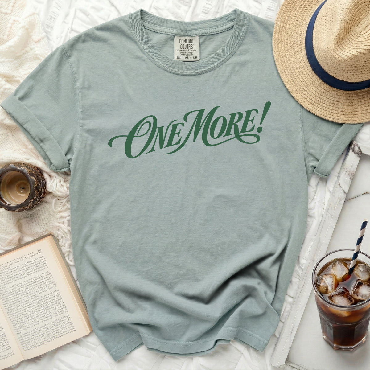 Green t-shirt with "ONE MORE!" graphic, book, hat, and drink.