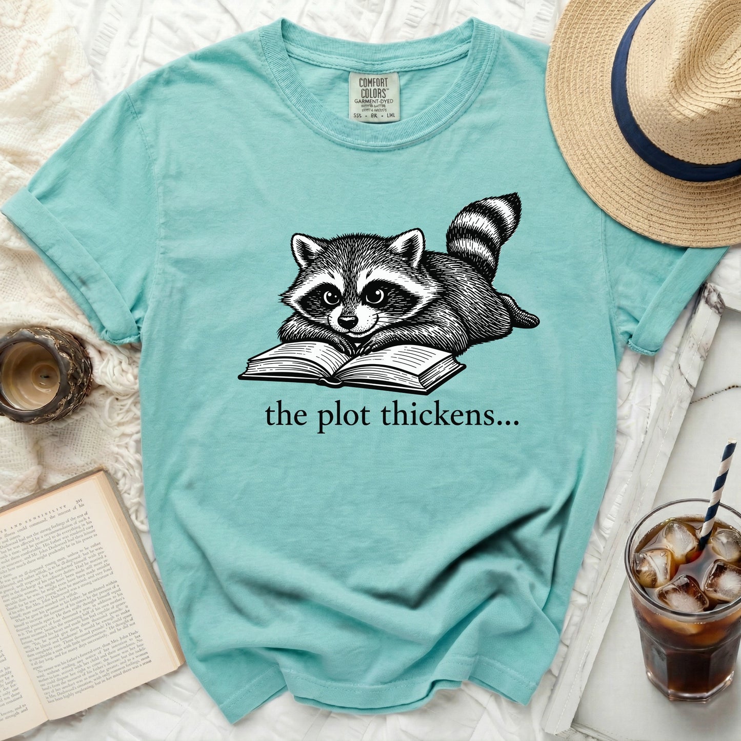 Teal t-shirt with a raccoon reading a book graphic and text "the plot thickens...".