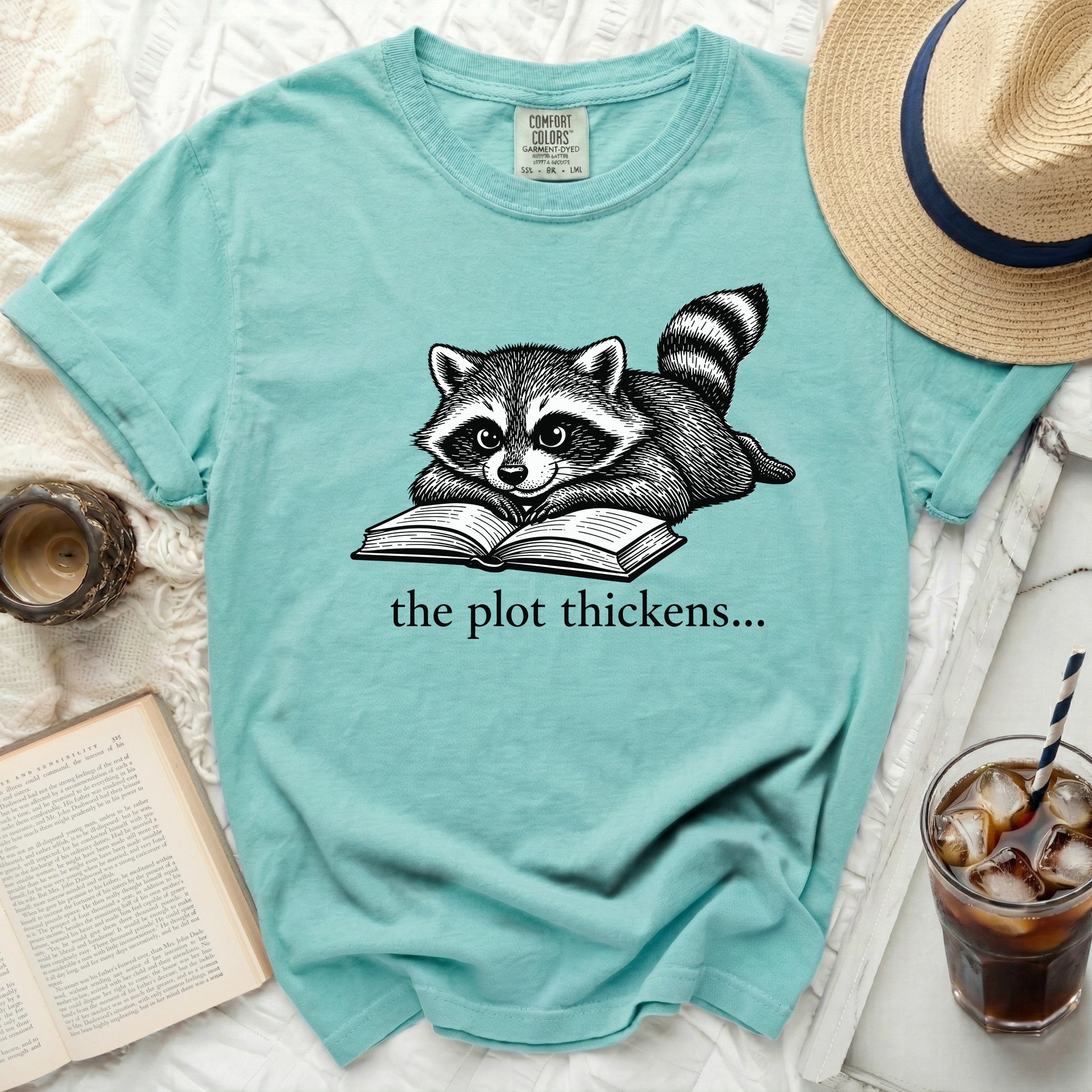 Teal t-shirt with a raccoon reading a book graphic and text "the plot thickens...".