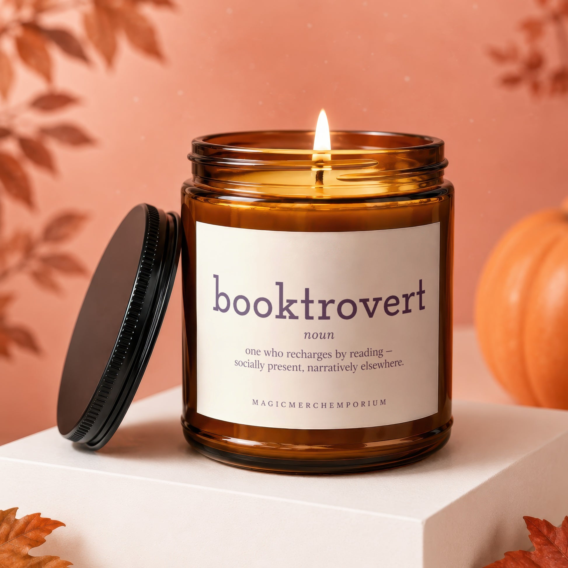 Amber glass candle with "booktrovert" definition label, lit flame, beside a black lid cap and pumpkin.