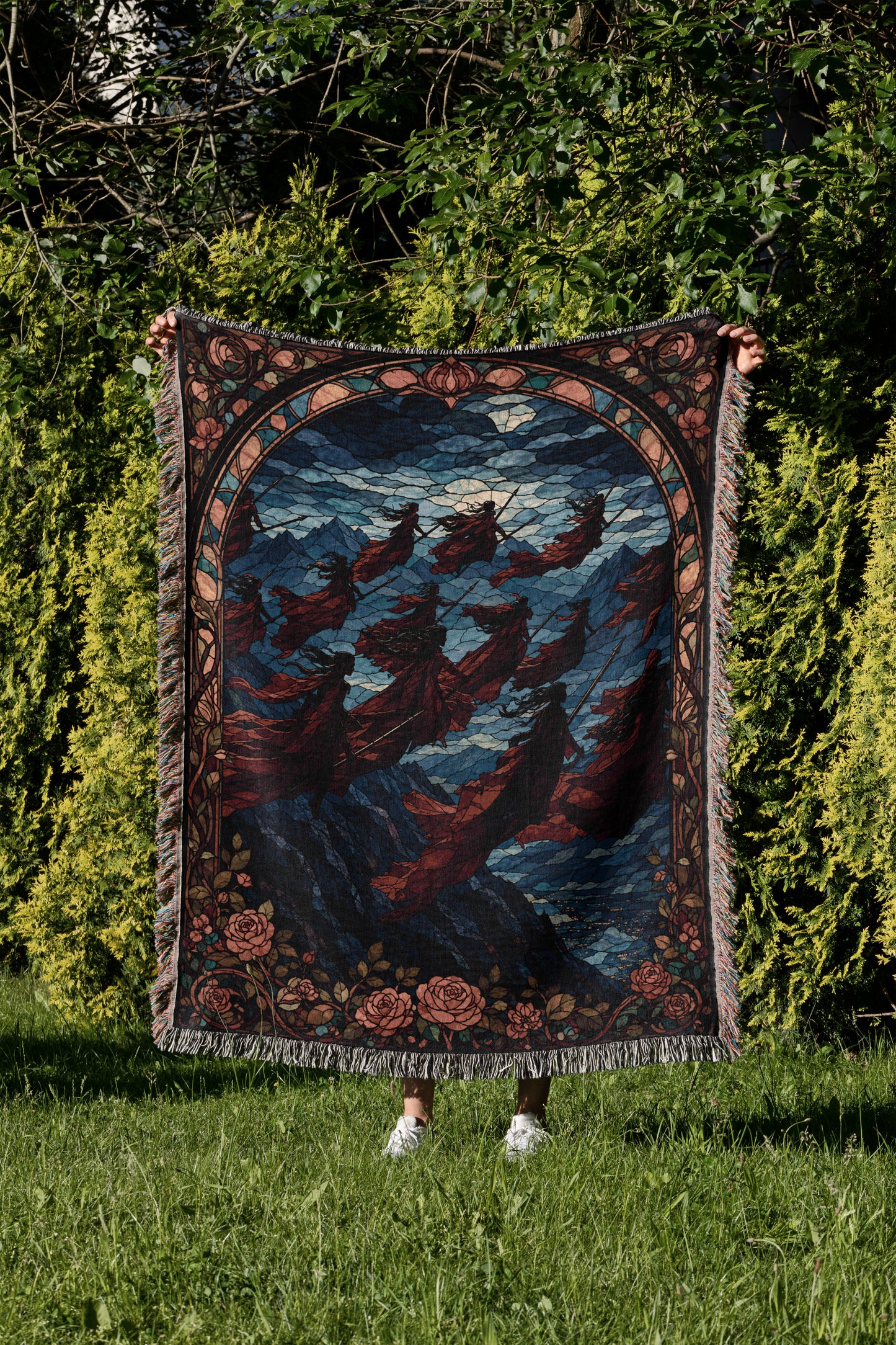 A person is holding up a large, colorful, and intricate rug with a floral design in a grassy area surrounded by trees.