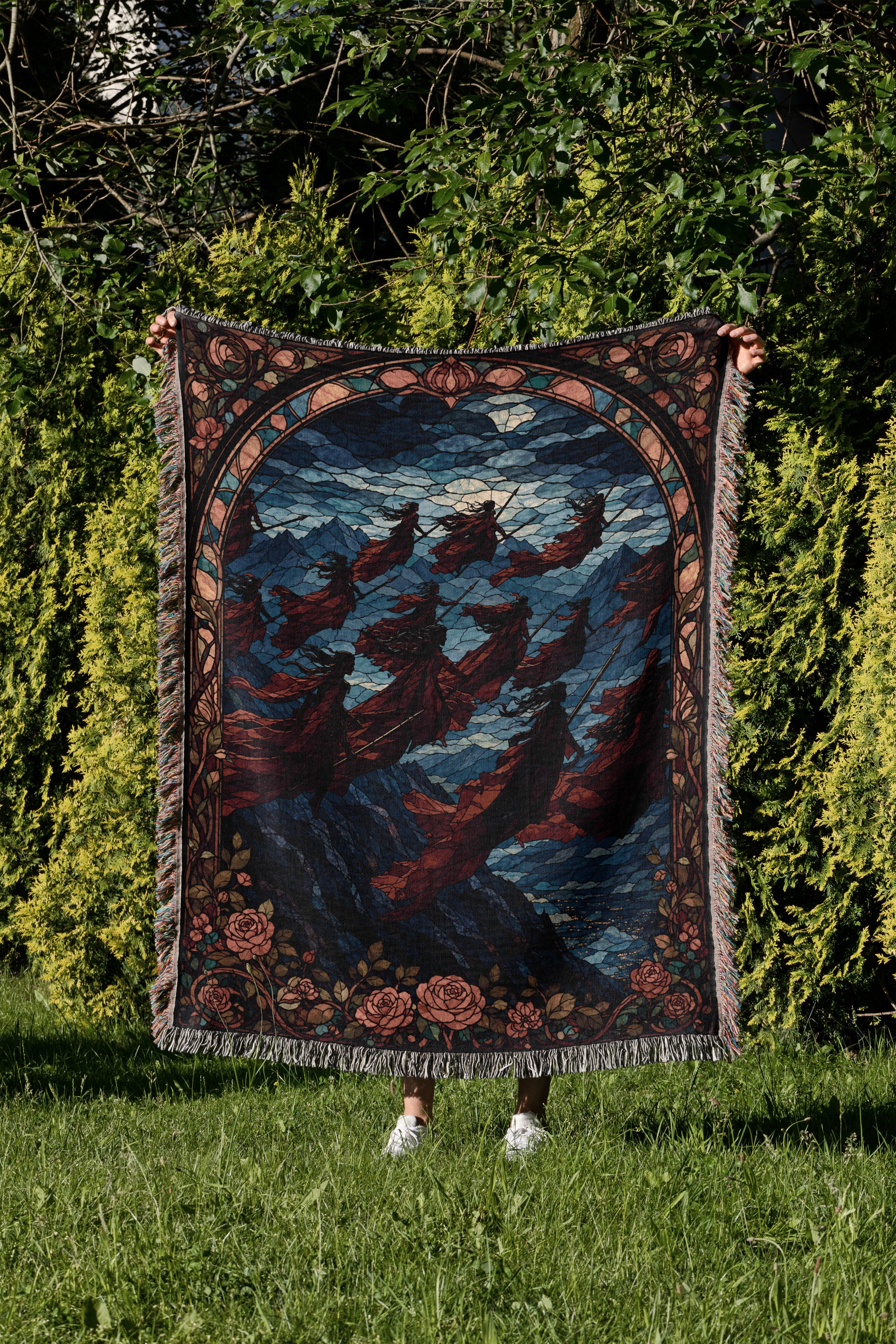 A person is holding up a large, colorful, and intricate rug with a floral design in a grassy area surrounded by trees.