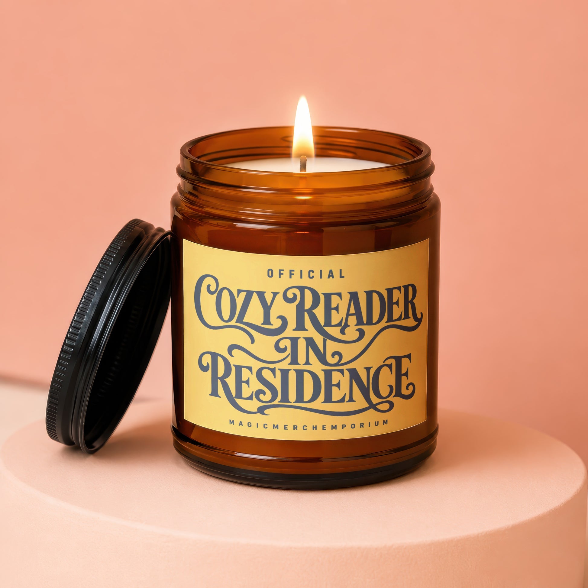 Amber jar candle with lit wick and lid off, reads "Official Cozy Reader In Residence"