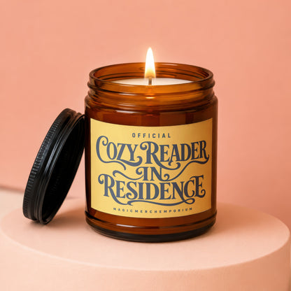 Amber jar candle with lit wick and lid off, reads "Official Cozy Reader In Residence"