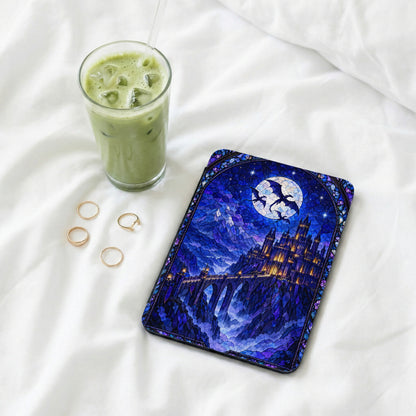 A Kindle case with a blue stained-glass castle design lies on a white bed alongside four gold rings and an iced matcha latte.