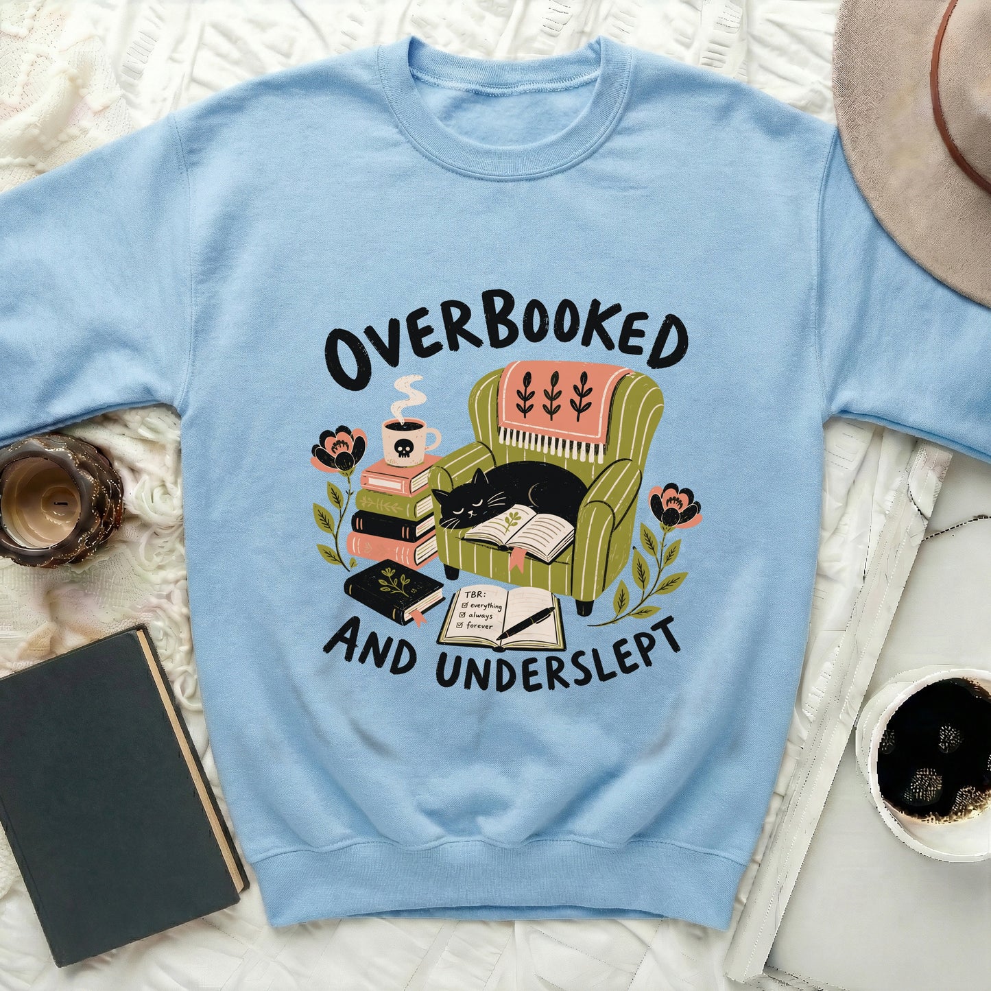 Light blue sweatshirt with "Overbooked and Underslept" text featuring a cat on an armchair.