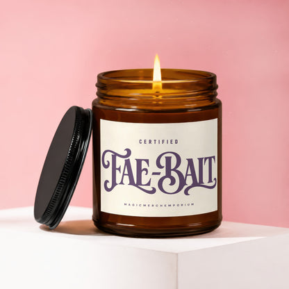 Glass candle, lit wick, "Fae-Bait" label, on a white surface against a pink background.