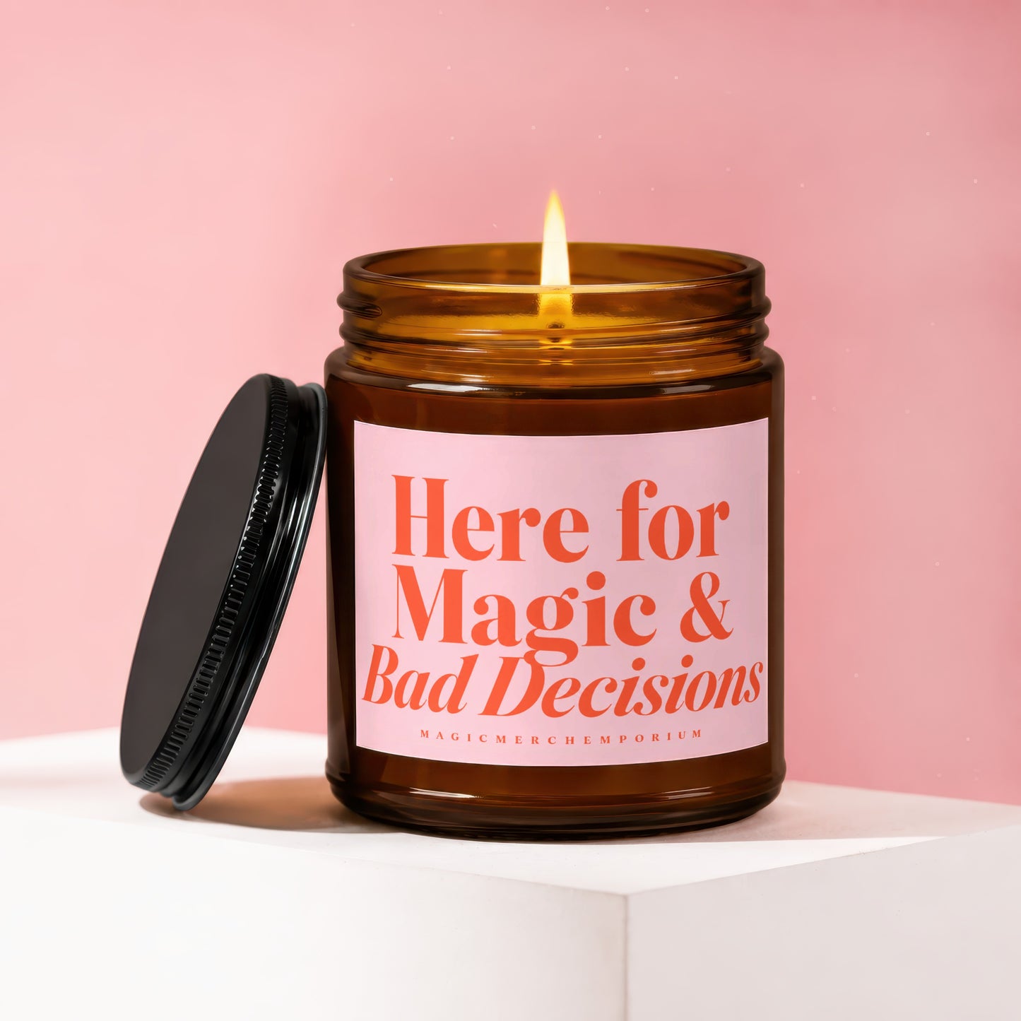 Amber glass candle with lit wick and pink label reading "Here for Magic & Bad Decisions".
