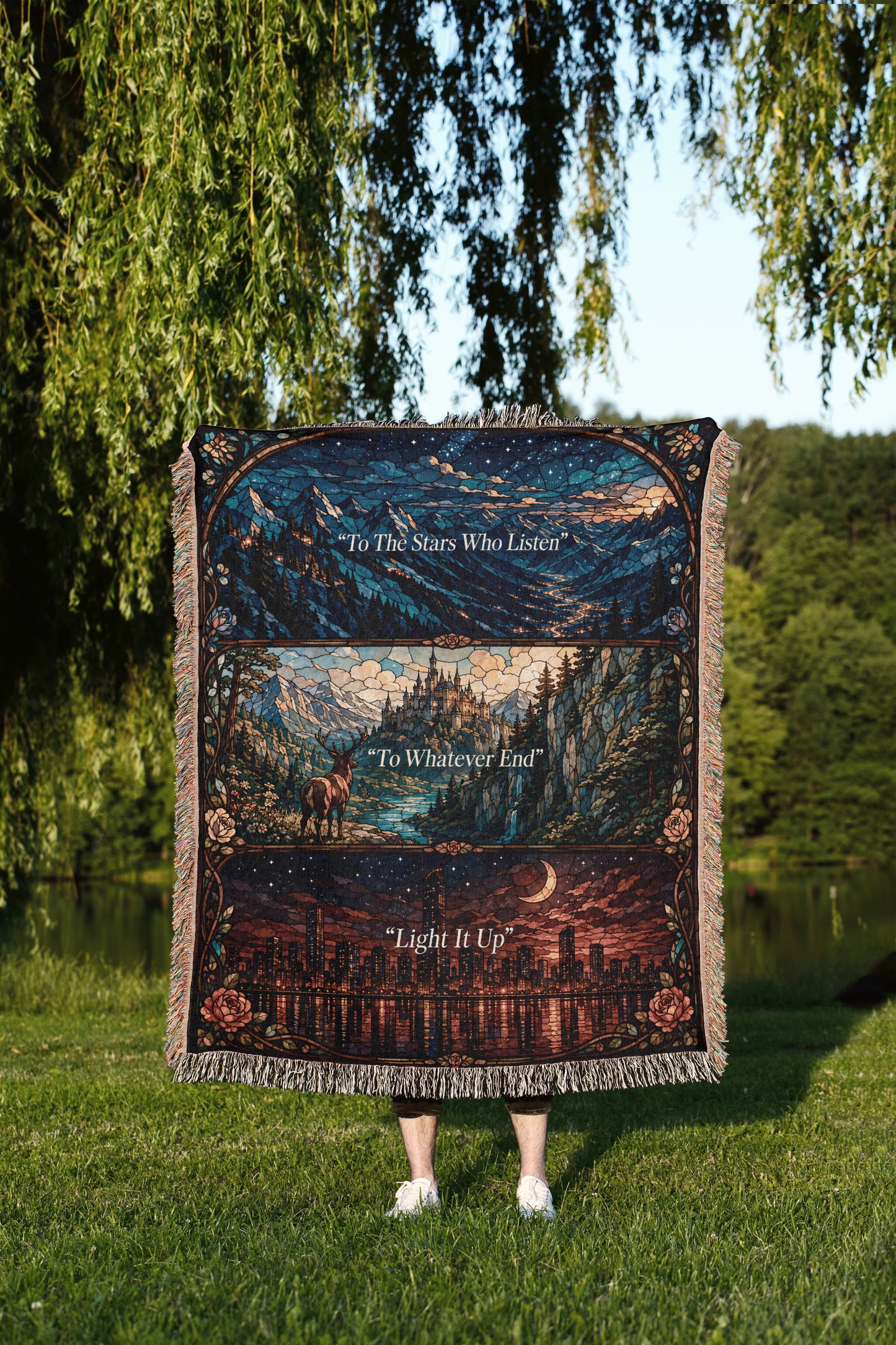 A person is standing in a grassy area, holding a large, colorful blanket that features a collage of images depicting a night sky, mountains, and a city skyline.