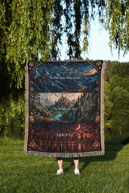 A person is standing in a grassy area, holding a large, colorful blanket that features a collage of images depicting a night sky, mountains, and a city skyline.