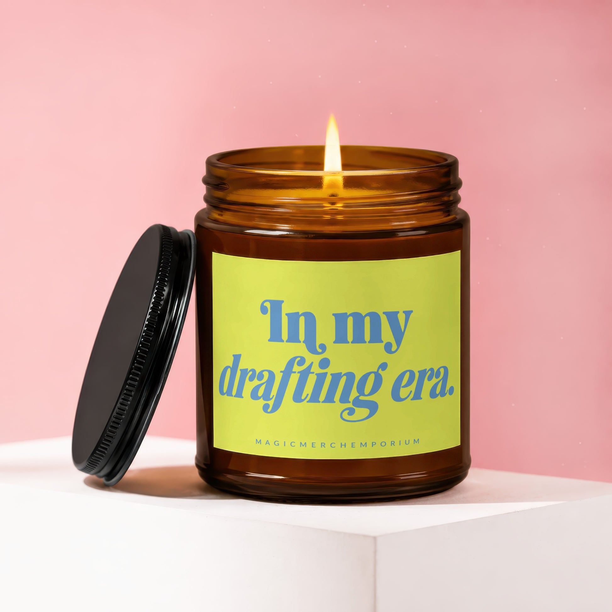 Lit candle in amber jar with black lid, reading "In my drafting era."