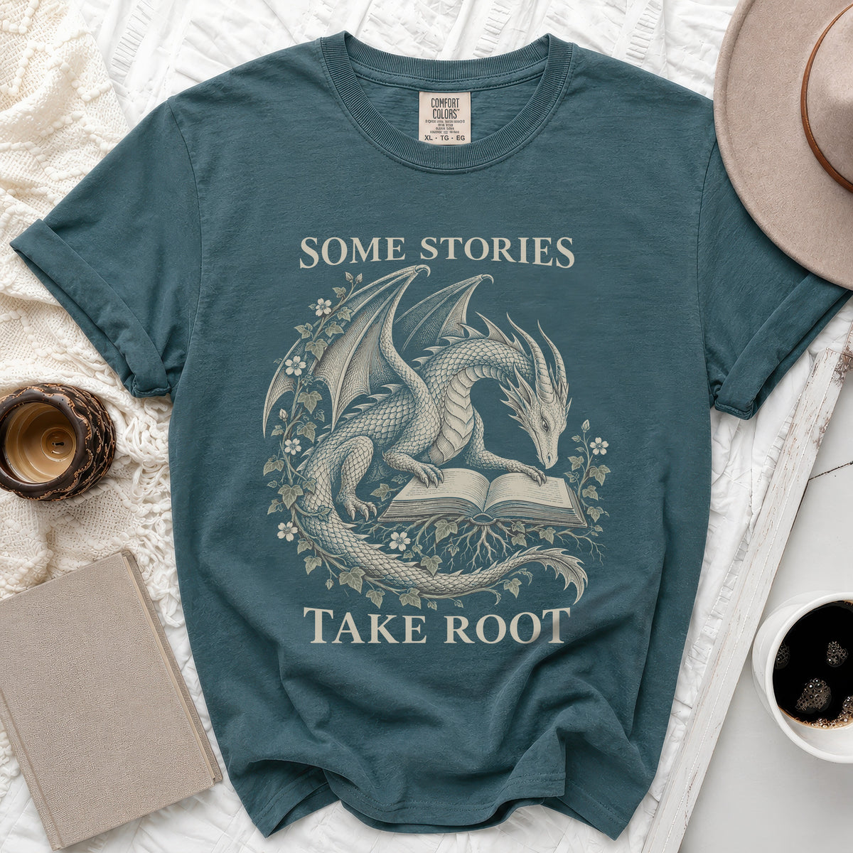 Teal t-shirt with dragon reading a book graphic and text "Some Stories Take Root".