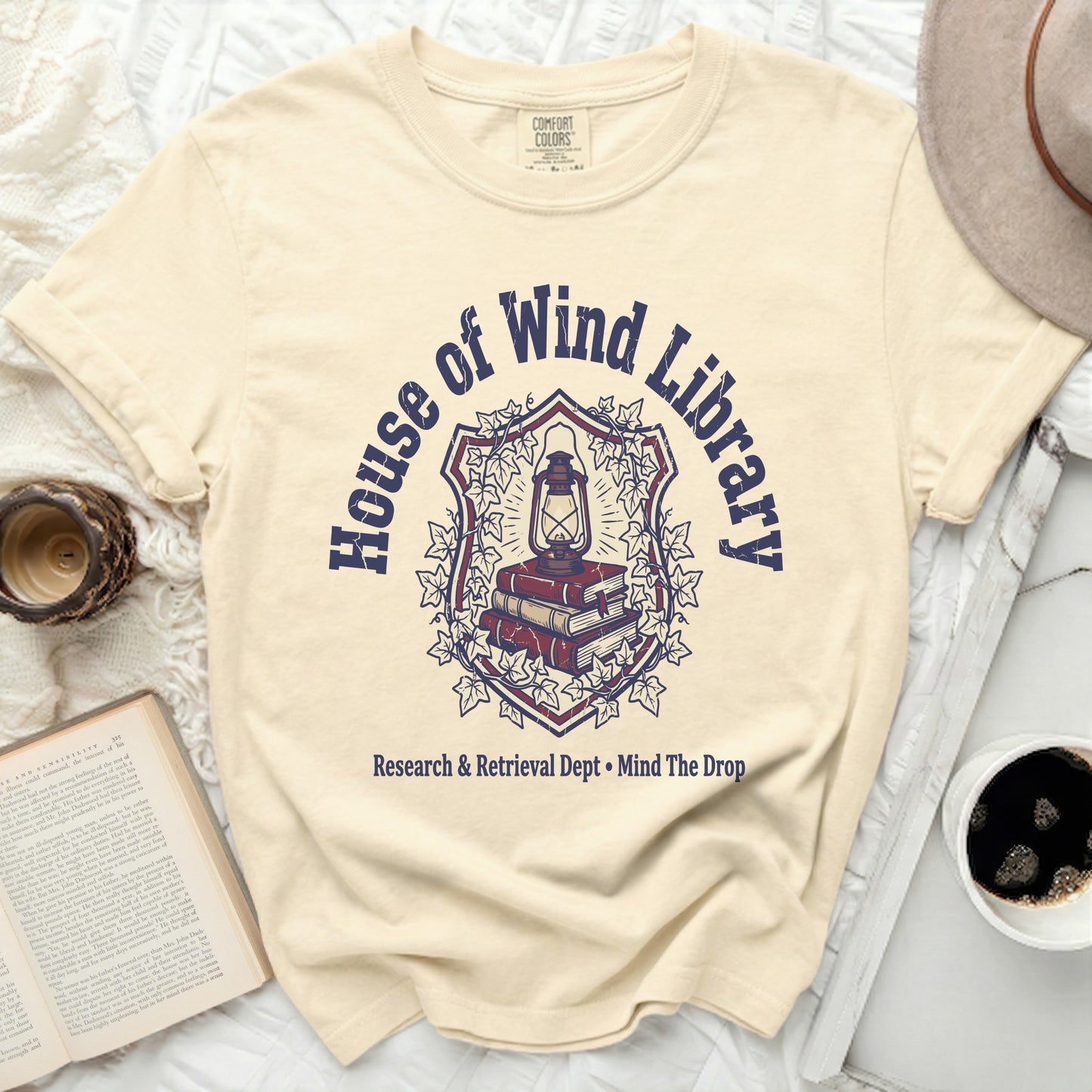 Cream t-shirt with "House of Wind Library" crest and book graphic.