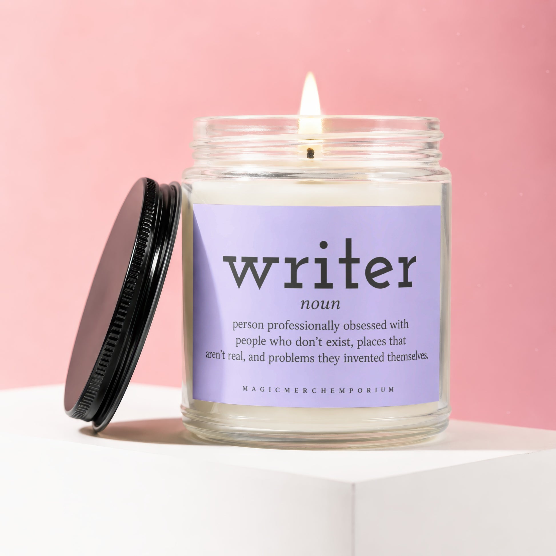 Candle with "writer" definition label, lit wick, and black lid on pink background.