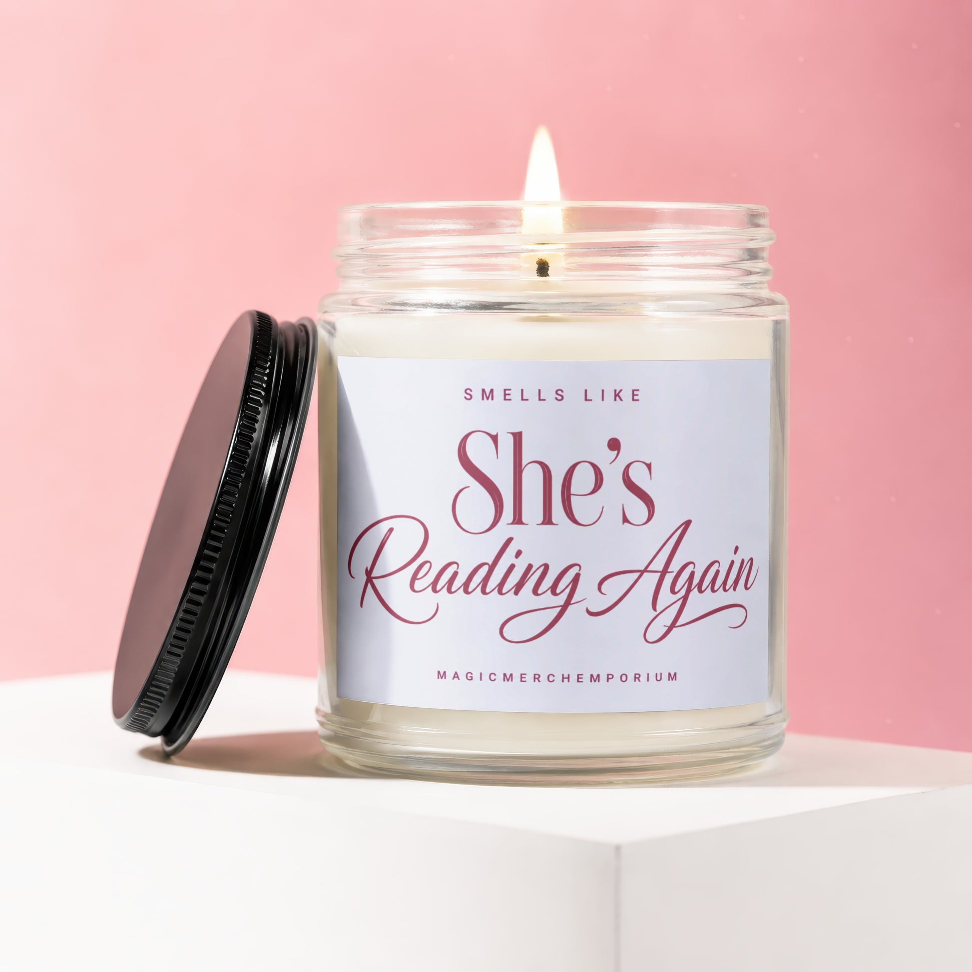 Candle with lit wick, "She's Reading Again" text, on a pink background.