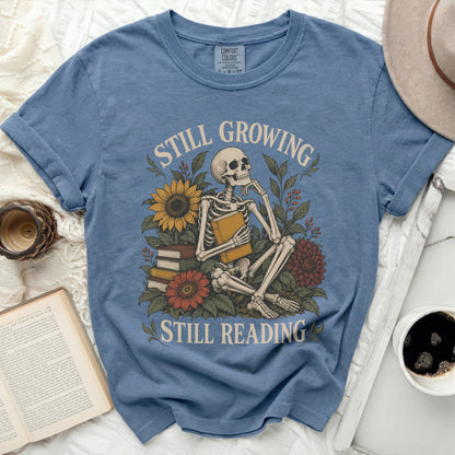 Skeleton with book and flowers, text "Still Growing Still Reading".