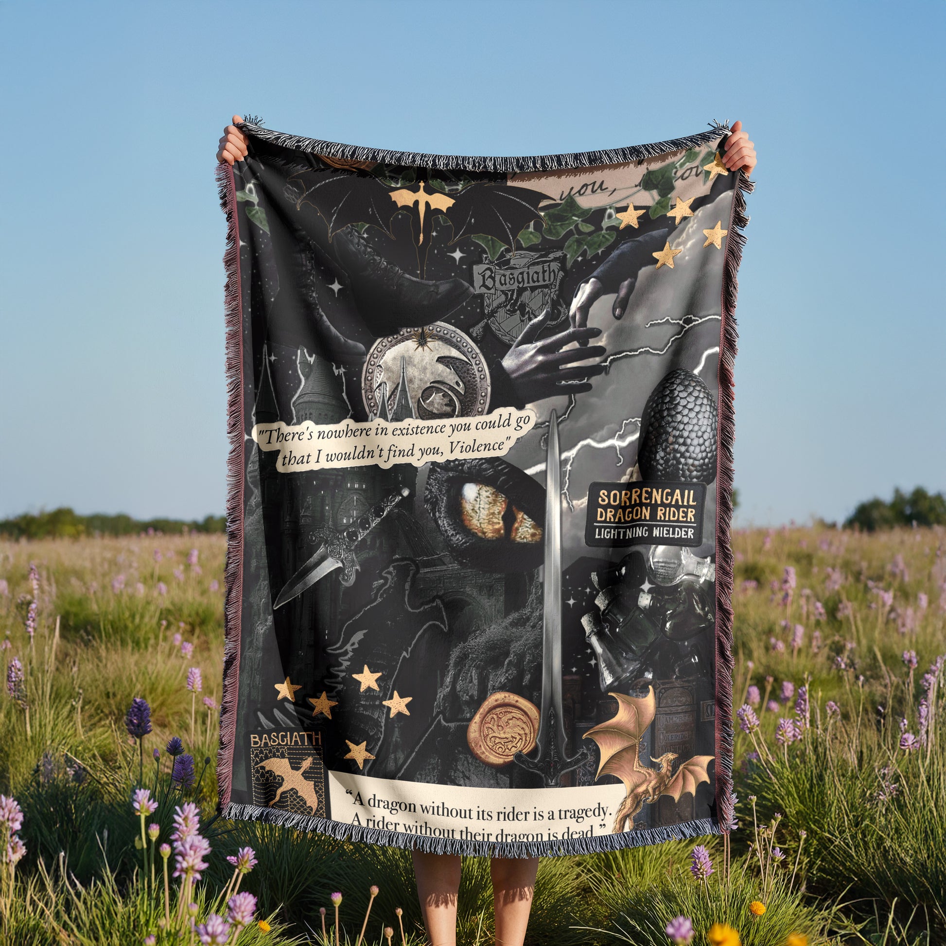 A person is holding up a large black and gold blanket with various images and text on it, standing in a field with purple flowers.