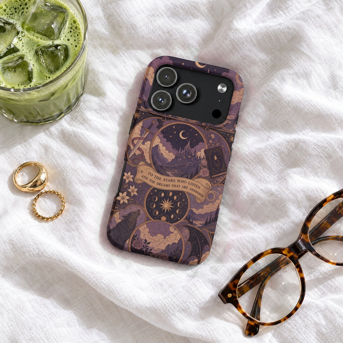 Purple phone case featuring celestial fantasy art, resting on a white blanket beside glasses, rings, and an iced matcha drink.