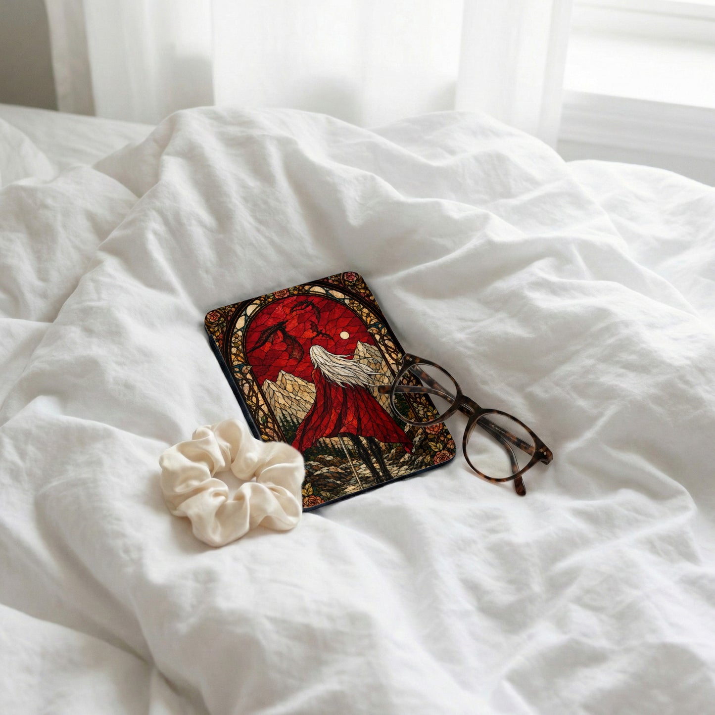 Book with stained glass cover art, glasses, and scrunchie on white bedding.