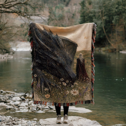 A person stands on a rock, holding a large tapestry with a dragon and a woman in a field in front of a river.