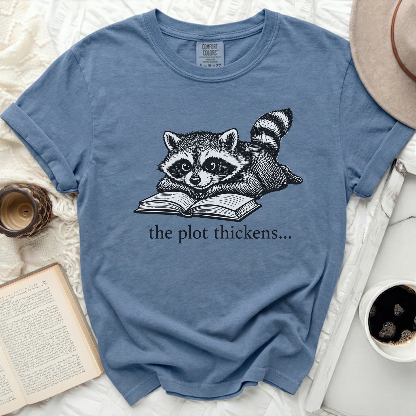 Blue t-shirt with a raccoon reading a book and text "the plot thickens..."