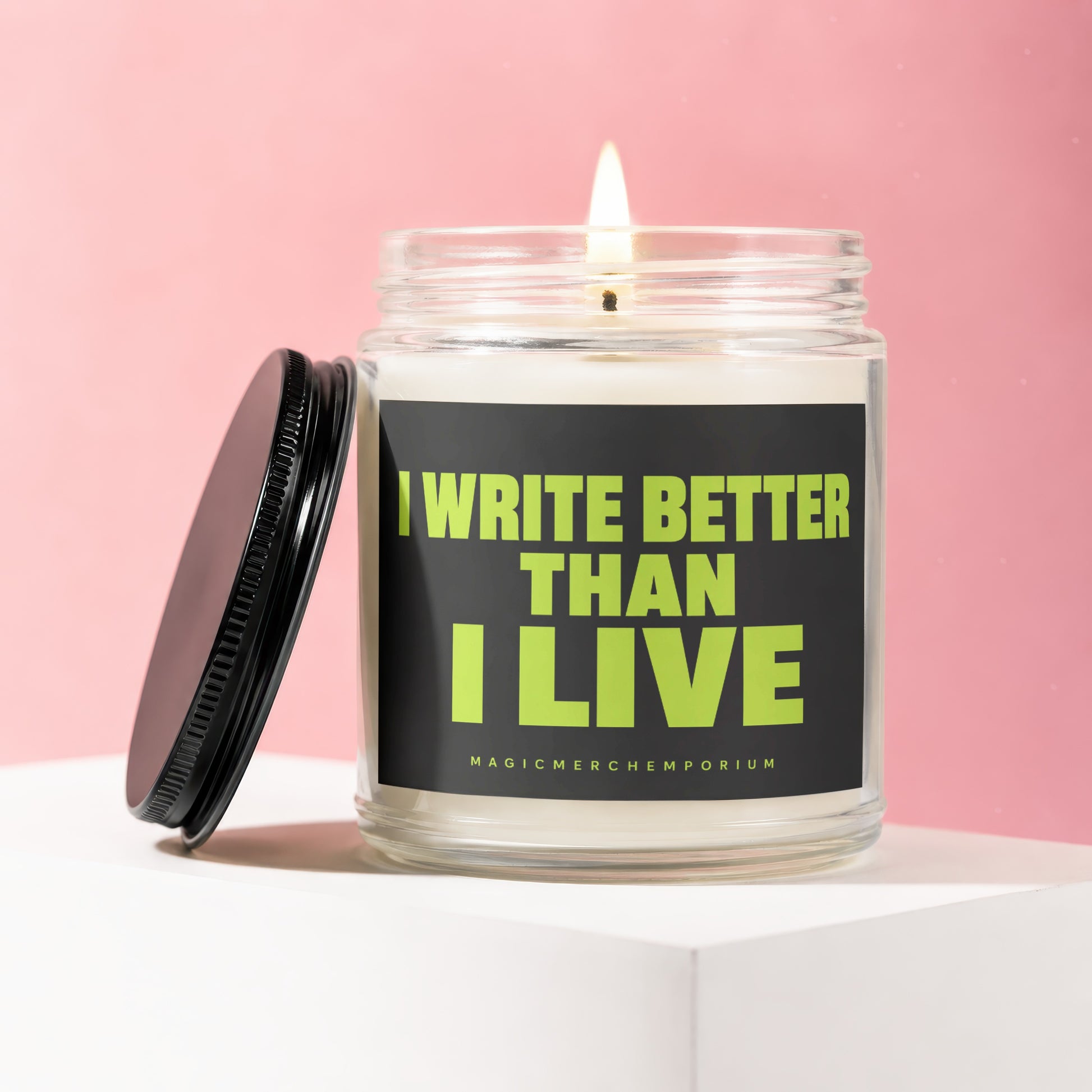 Glass candle with lit wick, black lid, and "I WRITE BETTER THAN I LIVE" text.