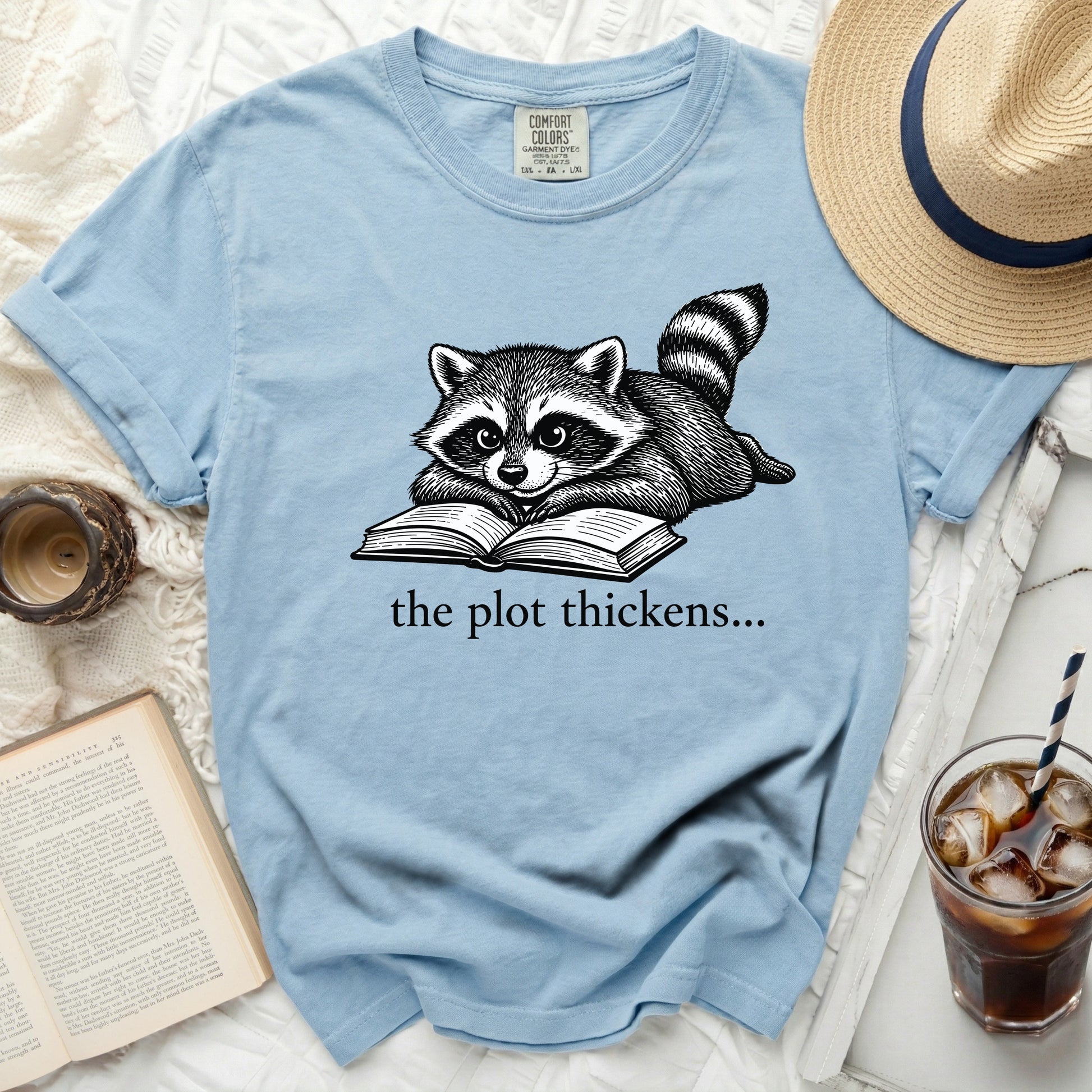 Light blue t-shirt with a graphic of a raccoon reading a book and the text "the plot thickens...".