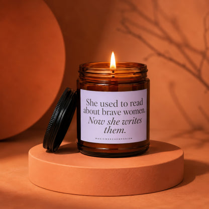 Amber jar candle with lit flame and lid off, saying "She used to read about brave women. Now she writes them."