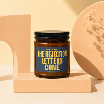 Amber jar candle with black lid reads "Light When: The Rejection Letters Come".
