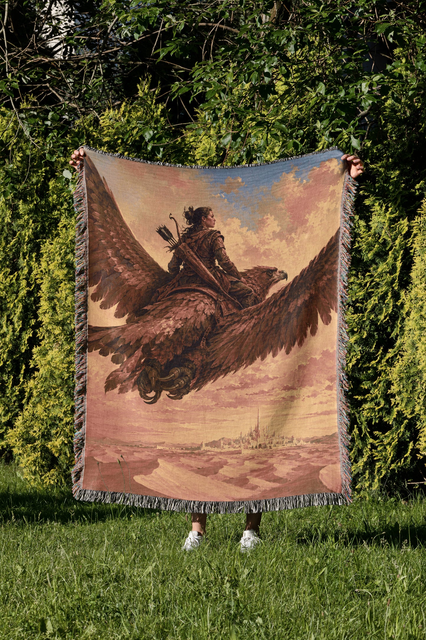 A person is standing in a grassy area, holding up a large tapestry or blanket that features a painting of a person riding a winged creature, possibly a dragon or eagle.