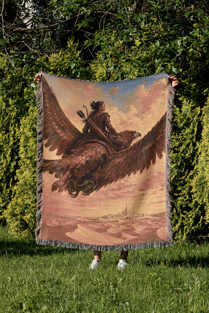 A person is standing in a grassy area, holding up a large tapestry or blanket that features a painting of a person riding a winged creature, possibly a dragon or eagle.