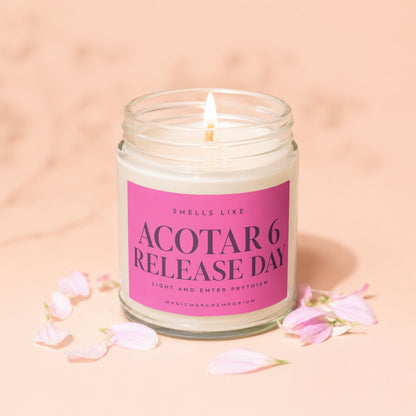 A lit candle in a glass jar with a pink label reading ACOTAR 6 RELEASE DAY, surrounded by light pink petals on a peach background.