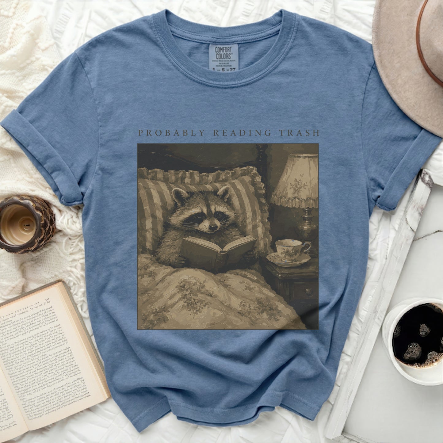 Blue t-shirt with raccoon reading in bed graphic and "PROBABLY READING TRASH" text.