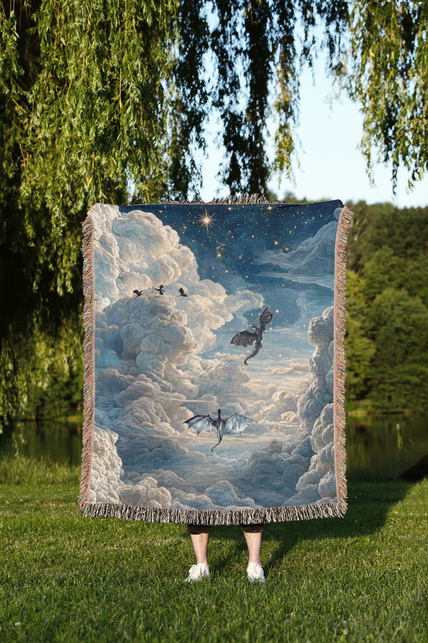 A person is standing in a grassy area, holding a large tapestry or blanket that features a painting of a night sky with clouds and two flying dragons.