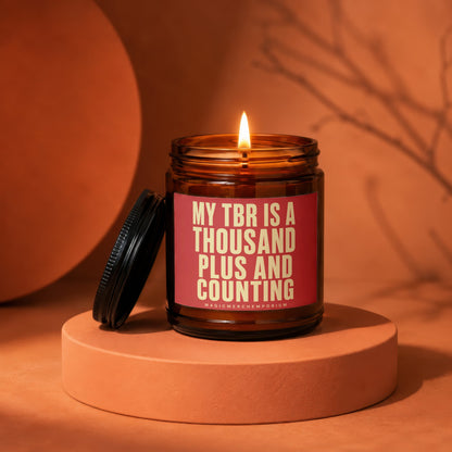 Brown glass candle with lit flame and red label reading "MY TBR IS A THOUSAND PLUS AND COUNTING".