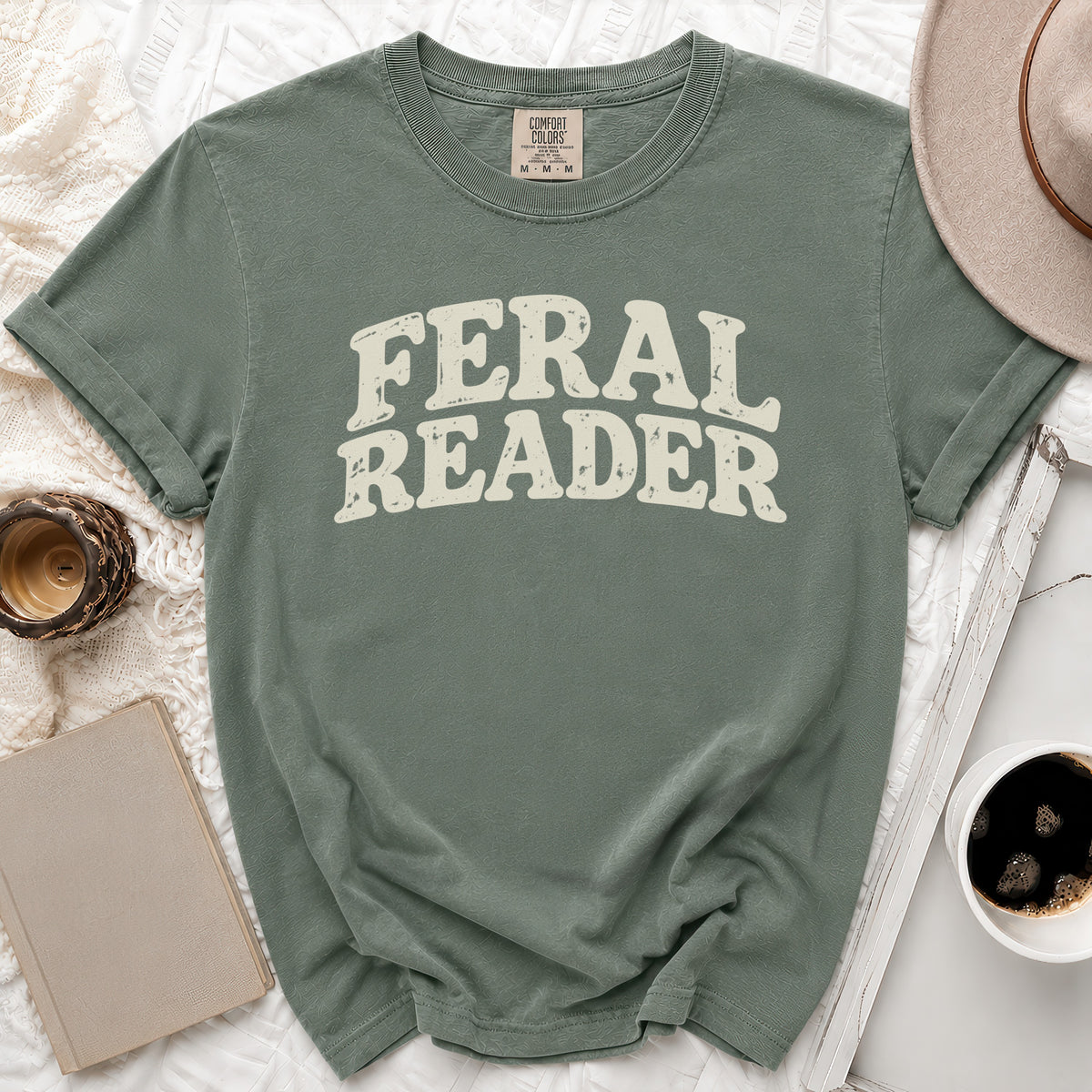 Green t-shirt with "FERAL READER" text, flat lay with hat, book, and coffee.